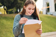 Attractive casual student girl joyfully opening envelope with exams results in park