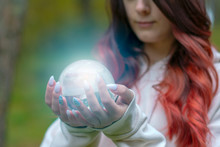 A Beautiful Young Witch Girl With Long Hair Is Holding A Mystical Magical, Glowing Crystal Ball In Her Hands In The Forest.