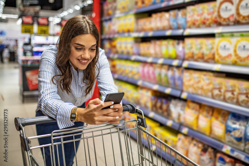 Fotografía Cropped image of girl leaning on shopping cart, using a mobile phone and smiling