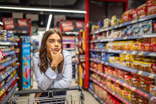 Young Woman Staying Confused In Grocery Store. Confused Woman Doesnt Know What To Buy. Young Woman With A Shopping Cart At Supermarket. Looking At Supermarket Shelf