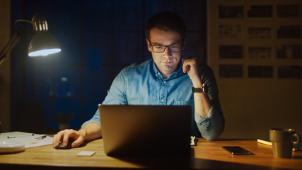 Poster - Professional Man Sitting at His Desk in Office Studio Working on a Laptop in the Evening. Man Analyzing Statistics.