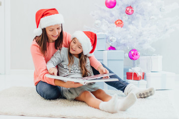 Pretty young mom reading an interesting book to her cute daughter near Christmas tree indoors.