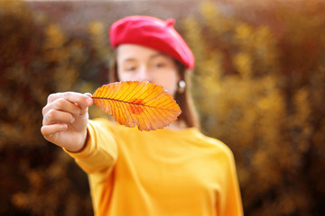 Wall Mural - autumn kiss. young girl in red bonnet holding yellow leaf with red lipstick kiss stamp on it outdoor
