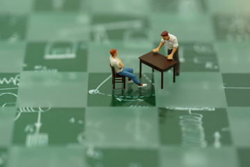 Miniature people sitting with table on Green chess board.