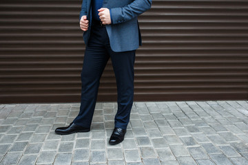 Wall Mural - Man's legs. Man wearing suit jacket, pants, blue shirt and black shoes standing near brown roller door on the city street. Details of classic elegant formal look. Trendy men's outfit. Street fashion.