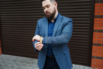 Wall Mural - Stylish brunette man with beard, wearing suit jacket and blue shirt, puts a watch on his hand against brown street wall. Details of classic elegant formal look. Trendy men's outfit. Street fashion.