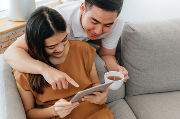 young asian love couple drinking coffee and using mobile tablet, watching movie online on sofa at home office, internet technology, social media, online booking, entertainment, shopping online concept