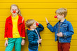 © Iryna - Sister with two brothers with red clown noses standing near yellow wall giving high five and smiles. Positive girl in red jacket looking at camera.