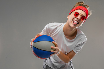 A cool young caucasian man with short red curly hair in white t-shirt, red glasses isolated on a grey background show a tongue and play basketball