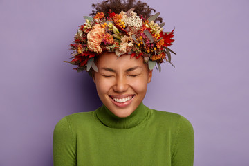 Happiness and season concept. Headshot of adorable Afro woman laughs out joyfully at funny joke, has fun with boyfriend, wears handmade autumnal wreath, smiles broadly, wears green turtleneck