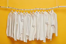 Group Of White Plain Clothes Hanging On Garment Rack Or Rail. Minimalistic Concept. Apparel For Women Isolated Over Yellow Background. Clothing Organization Or Storage. Row Of Sweaters And Tshirts