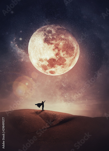 Fotografía Surreal scene with a woman hero silhouette with cape on the top of a hill raises hand up to the sky watching the full moon night