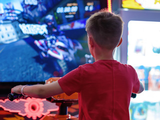 Wall Mural - Cute European boy in red t-shirt playing car racing on slot-machine. He enjoying his spare time. Back view.