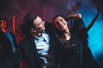 Watch my moves! Overjoyed young man in a smart outfit is spreading hands while dancing with his girlfriend, who is posing with her left hand over her head, smiling and laughing sincerely.