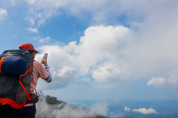 Wall Mural - A backpack woman standing on cliff and use mobile take photo for view in nature.Tourist enjoying fresh air.A woman enjoying free happiness in beautiful Thailand landscape.