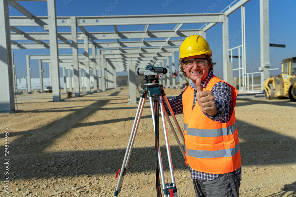 Smiling Construction Worker With Tacheometer Giving Thumb Up at a Construction Site - obrazy, fototapety, plakaty 