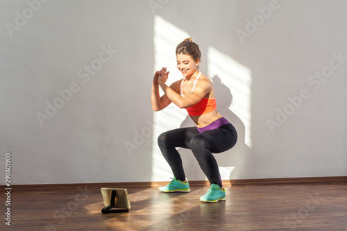 Fotomural Positive sportive woman with bun hairstyle and in tight sportswear doing squatting sit-up exercise while watching training video on tablet