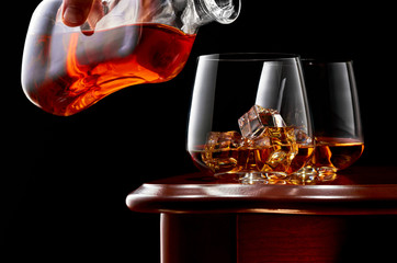 Two glasses of whiskey and ice on a wooden table in a low key. On top of the glass a man's hand with a bottle of whiskey.