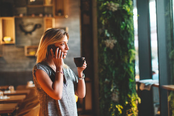 Beautiful young woman drinking coffee and talking on the phone in cafe and looking out the window