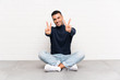 © luismolinero - Young handsome man sitting on the floor smiling and showing victory sign