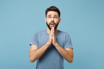 Wall Mural - Young pleading handsome man in casual clothes posing isolated on blue wall background, studio portrait. People sincere emotions lifestyle concept. Mock up copy space. Holding hands folded in pray.