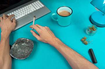 Person smoking a marijuana joint while working in a blue table with a computer and a coffee cup.