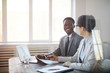 © Seventyfour - Portrait of two young entrepreneurs discussing project while sitting at meeting table behind glass wall, focus on smiling African businessman looking at partner, copy space