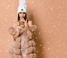 Woman In Party Dress And Fur Vest Is Scared With News She Has Got By Smartphone, Social Networks Under The Snow On Beige