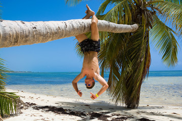 Poster - Caucasian teenage boy hanging on a palm tree on tropical beach in Sargasso sea, Punta Cana, Dominican Republic. Summer vacation concept