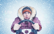 Frozen Man In Winter Clothes Drives A Car In A Snowstorm With His Hands On The Steering Wheel, Front View. 