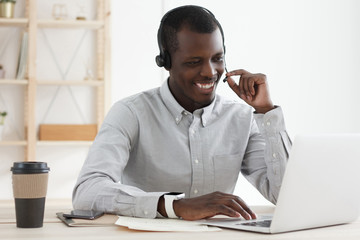 African man wearing headphones while working on laptop, answering calls from clients