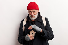 Old Man Wearing Street Clothes Holding House Made Of Cardboard, Dream About Shelter. Isolated Over White Background