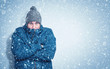 Frozen man in a blue jacket and hat stands against the wall, snow is falling around