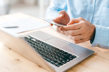 Poster - Young male employee scrolling or texting in smartphone over laptop keypad