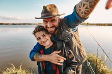 Father Hugs Son And Takes Selfie, Which Will Stand On Table At Home As Photo For Memory. They Have Fun. Background Lake.