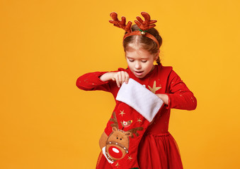 Sticker - happy funny child girl in red Christmas reindeer costume with gift surprise sock on yellow   background.