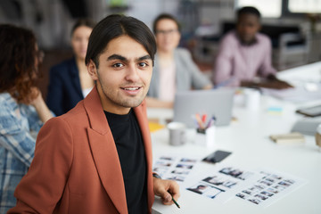 Wall Mural - Portrait of smiling Middle-Eastern man looking at camera while sitting at meeting table with creative team, copy space