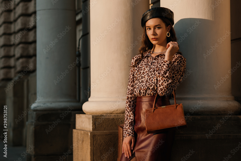 Outdoor fashion portrait of young elegant luxury woman wearing leopard print shirt, faux leather skirt, beret, holding small brown classic bag, handbag, posing in street Copy, empty space for text  - obrazy, fototapety, plakaty 