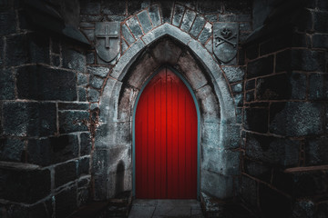 Scary pointy red wooden door in an old and wet stone wall building with cross, skull and bones at both sides. Concept mystery, death and danger.