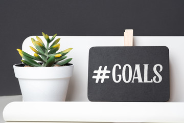 Poster - Goals on blackboard on pencil box and green plant in white pot on table and dark grey wall in office desk.business planning