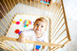 © Irina Schmidt - Beautiful little baby girl standing inside playpen. Cute adorable child playing with colorful toys. Home or nursery, safety for kids. Alone baby waiting for mom