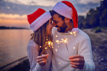 Overjoyed caucasian couple in love standing on coast near river and holding sparklers. Both are dressed in white sweaters and having santa hats on heads.