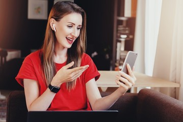 Wall Mural - Young woman sitting at home on sofa and using smartphone for communication with friends. Girl making video call with earphones and mobile phone. Digital technologies on wireless connected devices.