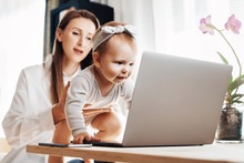 Young Mother With Baby Girl Sitting At Home In Front Of Computer And Watching Funny Videos For Children. Mom Showing Her Toddler Daughter Modern Digital Gadgets Using For Work And Education Online.