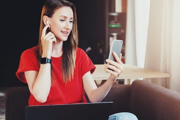 Wall Mural - Stylish young girl sitting with laptop on knees and listening to music with wireless earpads. Female in red shirt looking at screen of smartphone and streaming videos, watching stories, taking selfie.