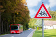 © wewi-creative - Attention, a red and white triangular road sign warns of cycling that can cross the road. Rural road at the forest with a red bus in the background.