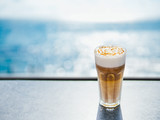 Hot latte macchiato coffee with caramel on tabletop with blue sea on background. Natural hard daylight. Copy space for text or design
