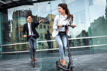 Two smiling business people driving electric scooter in front of modern business building going on work.