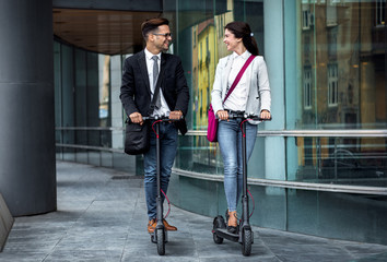 Two smiling business people driving electric scooter in front of modern business building going on work.