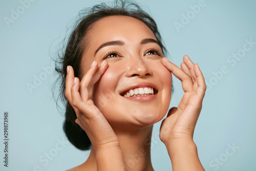 Zdjęcie: Skin care. Woman with beauty face touching healthy facial skin portrait. Beautiful smiling asian girl model with natural makeup touching glowing hydrated skin on blue background closeup Skin care. Woman with beauty face touching healthy facial skin portrait. Beautiful smiling asian girl model with natural makeup touching glowing hydrated skin on blue background closeup